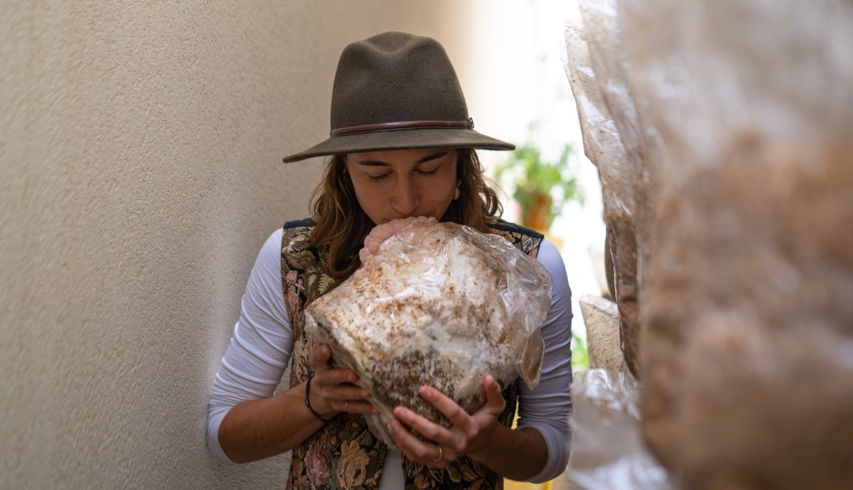 Happy woman smelling lion's mane mushroom grow kit fruiting block for easily growing mushrooms at home. The fruiting block is fully colonized and has large, premium lion's mane mushrooms starting to grow on it.