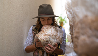 Happy woman smelling lion's mane mushroom grow kit fruiting block for  easily growing mushrooms at home. The fruiting block is fully colonized and has large, premium lion's mane mushrooms starting to grow on it.
