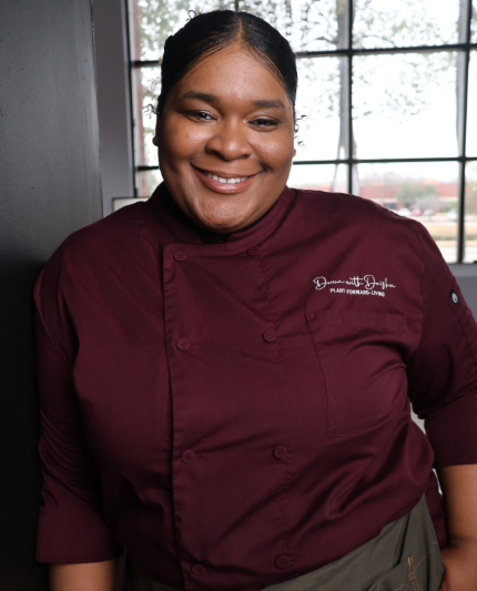 Close up of Daishu The Mushroom Queen, smiling in a red chef uniform with her name on it. 