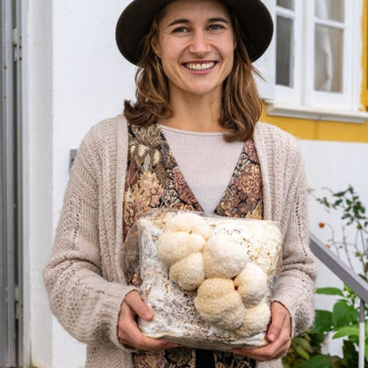 Smiling woman holding lion's mane mushroom grow kit fruiting block with large white lion's mane mushrooms growing on it.